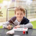 Little 7 year old boy paints greeting card for Mom on Mother's Day with the inscription "Mother's Day". Outdoors. Mother's Day