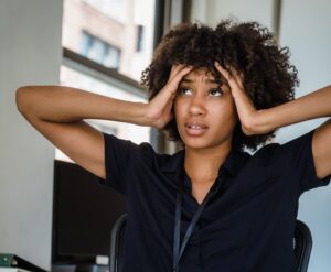 Image of a women in an office setting. The closeup photo features her face with a frustrated expression and her hands on framing her face.